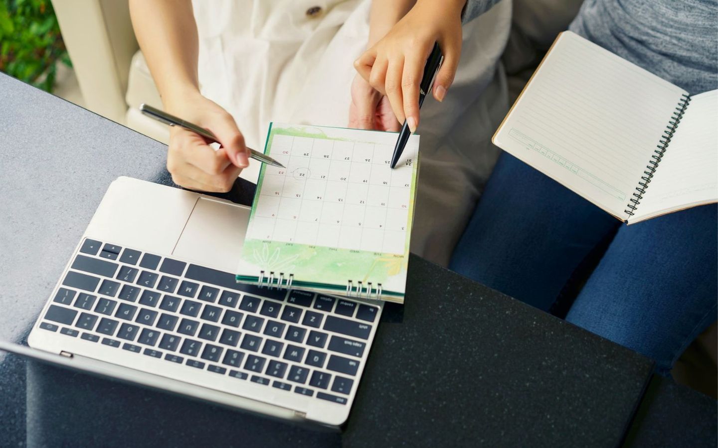 Ein Kalendar liegt auf dem Schreibtisch, zwei MitarbeiterInnen zeigen mit Ihren Stiften auf ein Datum